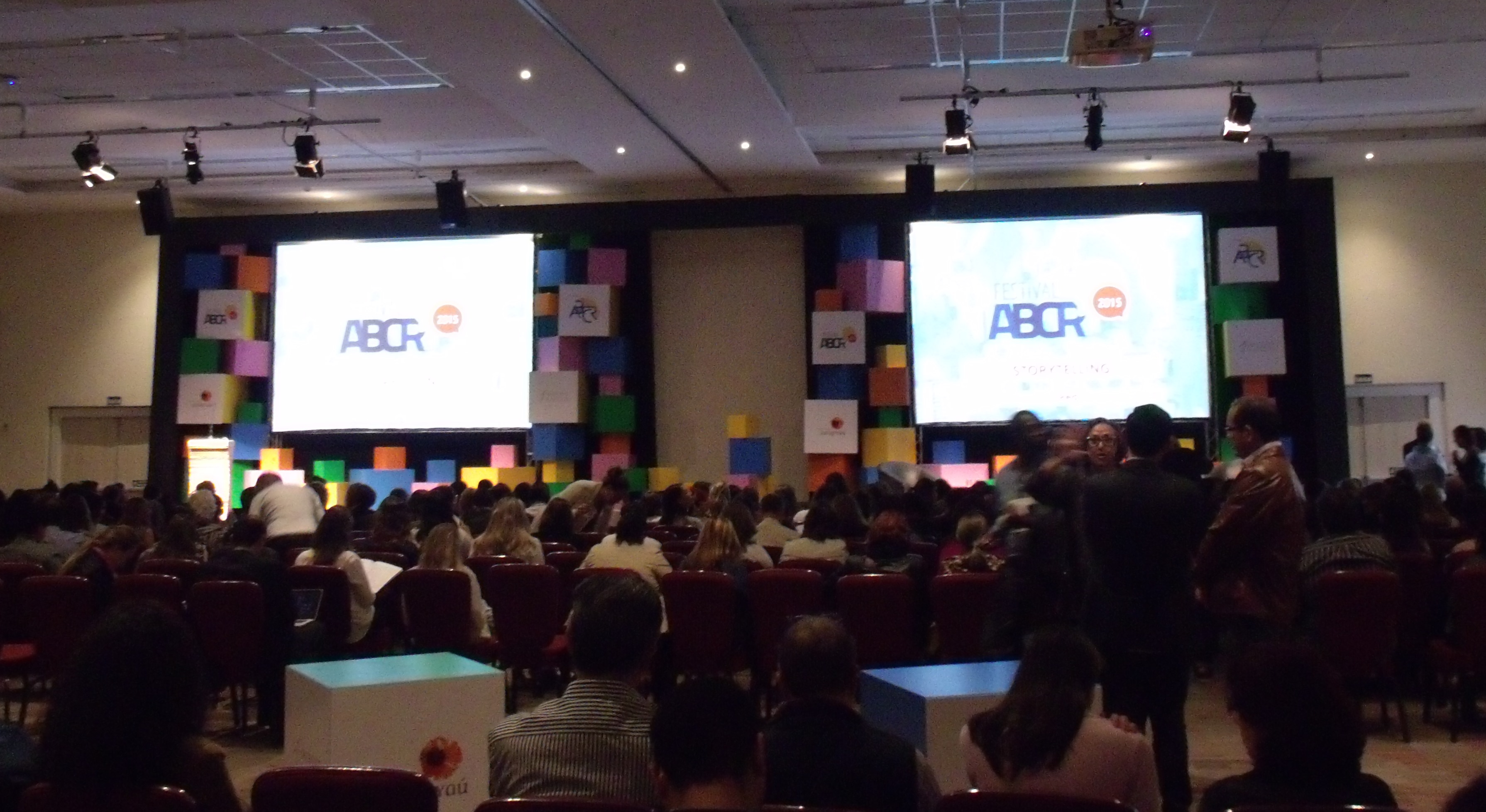 Foto tirada em um salão de eventos com muitas pessoas sentadas, de costas, e observando dois telões ao fundo