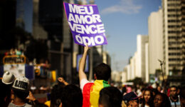 Foto de um homem segurando uma placa com fundo roxo e os dizeres em branco "Meu amor vence o ódio", durante parada LGBT de São Paulo.