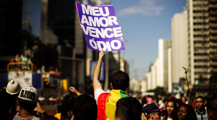 Foto de um homem segurando uma placa com fundo roxo e os dizeres em branco "Meu amor vence o ódio", durante parada LGBT de São Paulo.