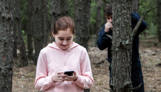A maniac watches from behind a tree in the forest for a teenage girl in a tracksuit with a mobile phone in her hands. The concept of child abduction.