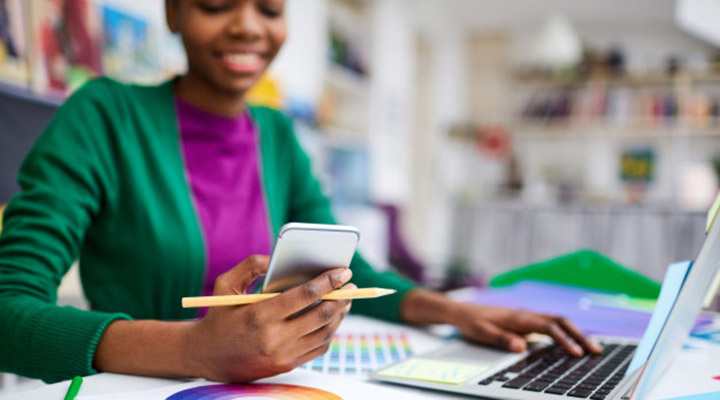 Mulher negra (em segundo plano), segura e sorri para smartphone (em primeiro plano), enquanto também interage com teclado de laptop.