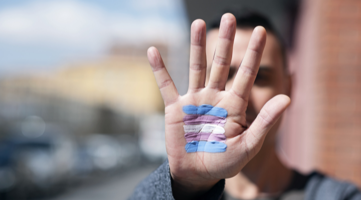 Foto tirada com o foco em primeiro plano da palma da mão de um homem aberta, mostrando uma pintura da bandeira trans. 
