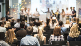 Imagem de auditório, com jovens sobre palco recebendo prêmio, em segundo plano; Em primeiro plano, uma plateia sentada assiste à premiação.