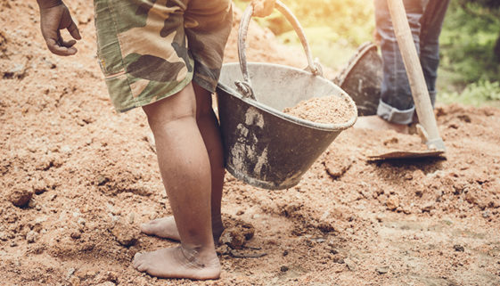 Foto dos pés descalços e parte das pernas de uma criança que carrega um balde com terra. Ao fundo, estão o que parecem ser as pernas de um adulto manipulando uma enxada. Está de dia.