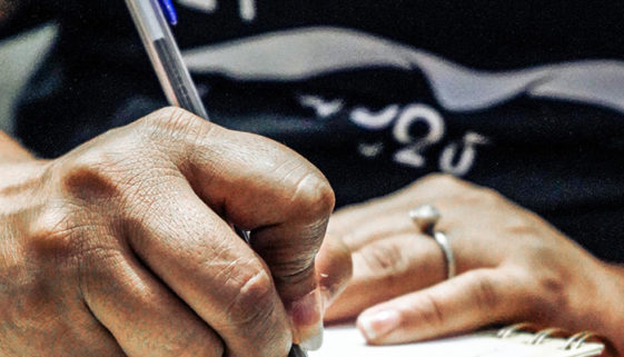 Mão de mulher negra escreve, com caneta de tinta azul, em caderno de espiral.