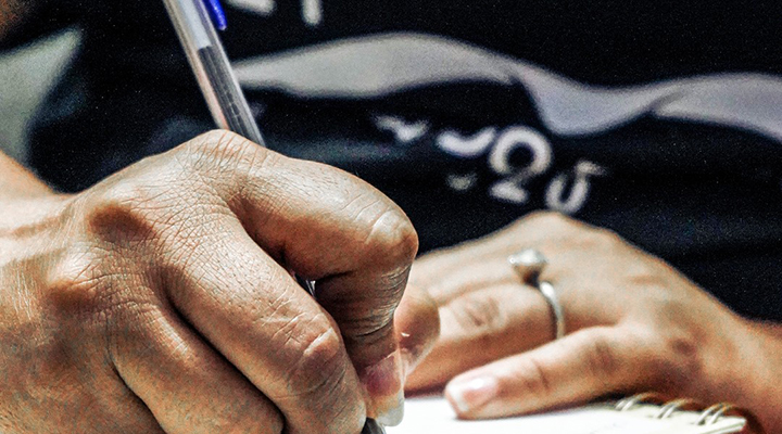 Mão de mulher negra escreve, com caneta de tinta azul, em caderno de espiral.