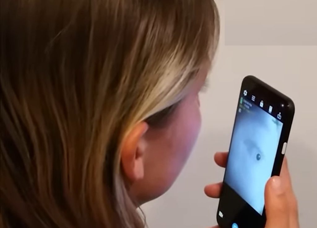 Foto do perfil direito do rosto de uma mulher branca, de cabelos louros, segurando um celular preto na frente do rosto. Na tela do celular é possível ver imagem do olho da moça., em tempo real.