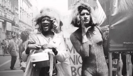 Foto em preto e branco tirada durante manifestação por direitos da comunidade LGBTQIA  em 1970. Marsha P. Johnson e Sylvia Rivera, ativistas transexuais da época estão em primeiro plano.