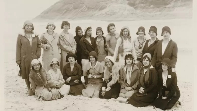 Foto em preto e branco das participantes do II Congresso Internacional Feminista no Recreio dos Bandeirantes (RJ). Onze moças estão de pé e oito estão apoiadas de joelhos na areia.