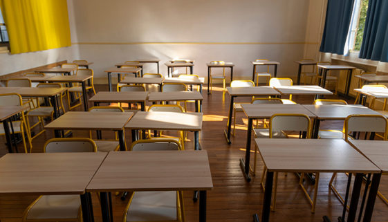 Fileiras de carteiras duplas com tampos de madeira estão dispostas em sala de aula vazia, iluminada pelas frestas das cortinas penduradas nas janelas do canto direito da imagem.