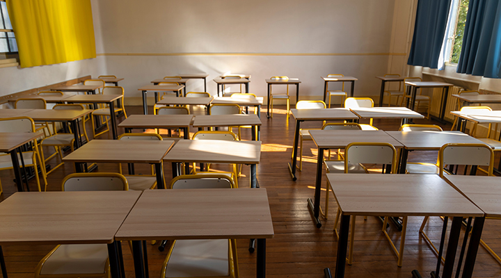 Fileiras de carteiras duplas com tampos de madeira estão dispostas em sala de aula vazia, iluminada pelas frestas das cortinas penduradas nas janelas do canto direito da imagem.
