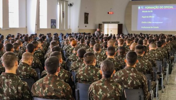 Foto tiradas das costas de alunos da Escola Preparatória de Cadetes do Exército, sentados em fileiras dispostas em um galpão e tomando aula.
