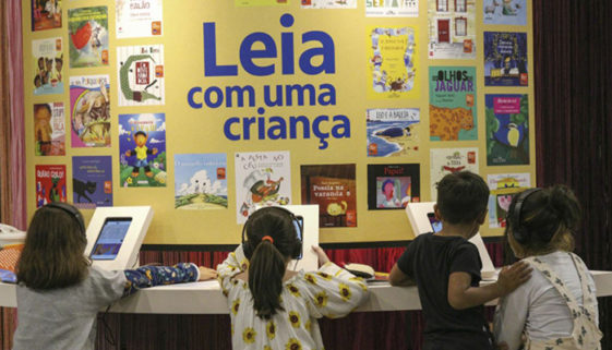 Foto tirada das costas de quatro crianças interagindo com totens digitais em uma bancada que circunde um quadro amarelo com vários pequenos cartazes espalhados em volta da frase 