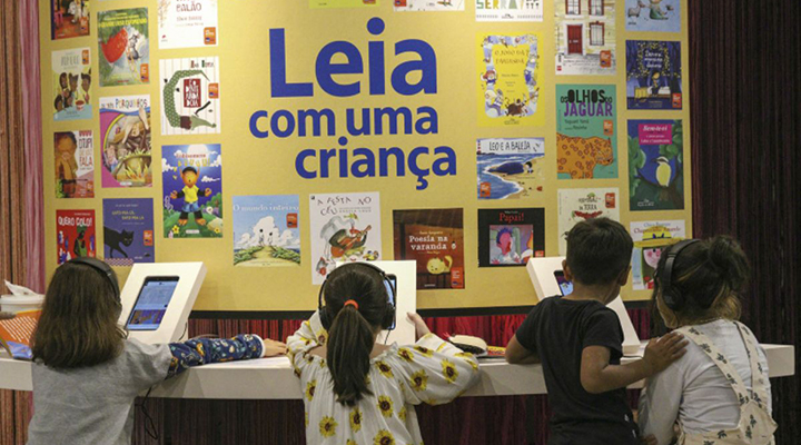 Foto tirada das costas de quatro crianças interagindo com totens digitais em uma bancada que circunde um quadro amarelo com vários pequenos cartazes espalhados em volta da frase "Leia com uma criança", de cor azul.