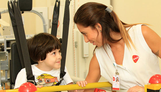Foto de mulher e criança se entreolhando com ternura. A criança está em um aparelho ortopédico sendo assistida pela adulta.