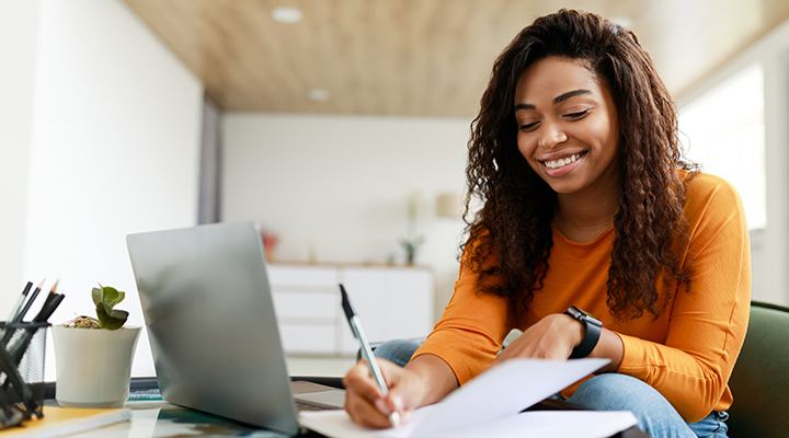 Sorrindo, jovem negra está sentada na mesa, trabalhando no laptop e escrevendo em papel branco.