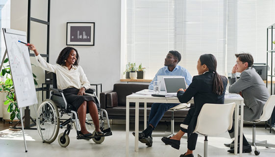 Empresária negra em cadeira de rodas apontando para flipchart e apresentando seu relatório aos colegas durante o trabalho em equipe no escritório