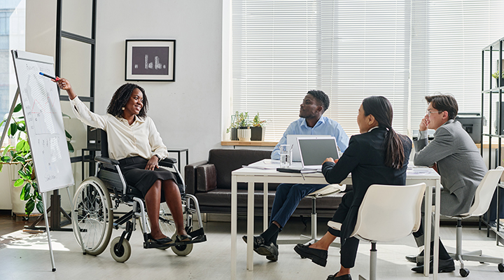 Empresária negra em cadeira de rodas apontando para flipchart e apresentando seu relatório aos colegas durante o trabalho em equipe no escritório