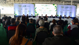 Foto de plateia à frente de mesa comprida com palestrantes. Ao fundo estão três telões expositores.