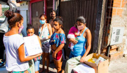 Foto de quatro mulheres interagindo com uma quinta que segura uma caixa nas mãos. Uma delas carrega uma criança no colo. Um homem aparece ao fundo. O dia está ensolarado.