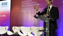 Foto de homem branco vestindo terno e gravata pretos, por cima de camisa branca, palestrando em púlpito, à frente de telão com fundo rosa e o título em branco e em letras garrafais: Seminário Internacional SESI de Educação. 
