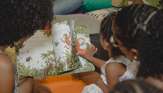Foto das costas de duas crianças e uma mulher negras interagindo com um livro cheio de desenhos.