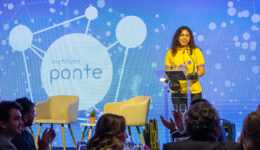 Foto de garota vestindo blusa amarelo, sobre um palco, falando em púlpito para uma plateia. Um telão com fundo azul e alguns círculos brancos preenchem o fundo da foto.