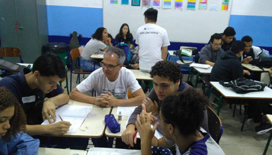 Foto de alunos e professores sentados em grupos em sala de aula.