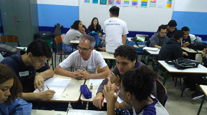 Foto de alunos e professores sentados em grupos em sala de aula.