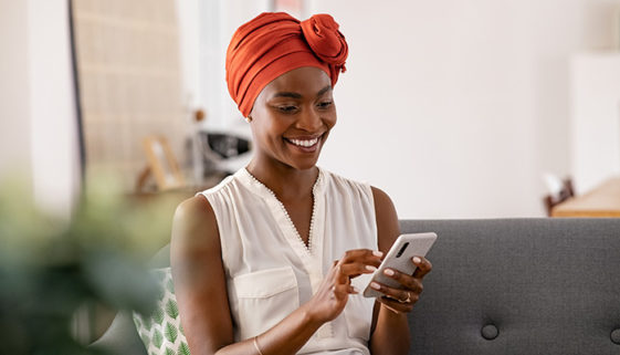  Mulher negra, de meia idade, com turbante tradicional. Está sentada no sofá, e sorrindo enquanto usa smartphone. 