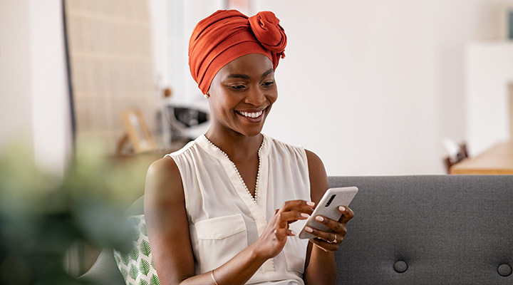  Mulher negra, de meia idade, com turbante tradicional. Está sentada no sofá, e sorrindo enquanto usa smartphone. 