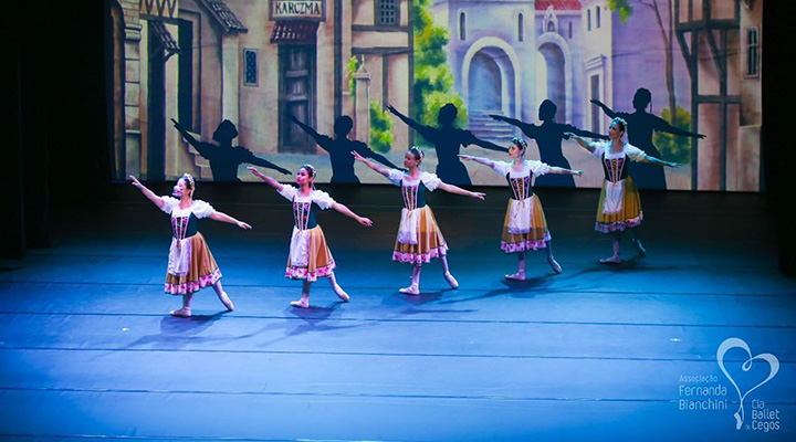 Foto tirada de bailarinas em posição sincronizada com os braços abertos. Elas estão se apresentando em um palco com piso de madeira e fundo que reproduz uma espécie de vilarejo.