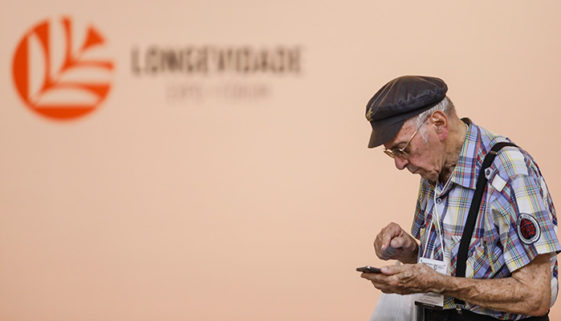 Foto de homem idoso usando boina, camisa xadrez, e calça com suspensório, interagindo com um celular. Ao fundo, uma parede cor salmão leve o logo e o nome do evento aparecem desfocados.