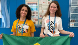 As duas garotas estão sorrindo para a foto enquanto seguram, juntas, uma bandeira do Brasil
