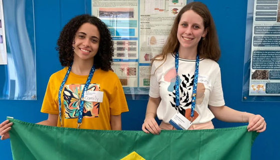 As duas garotas estão sorrindo para a foto enquanto seguram, juntas, uma bandeira do Brasil