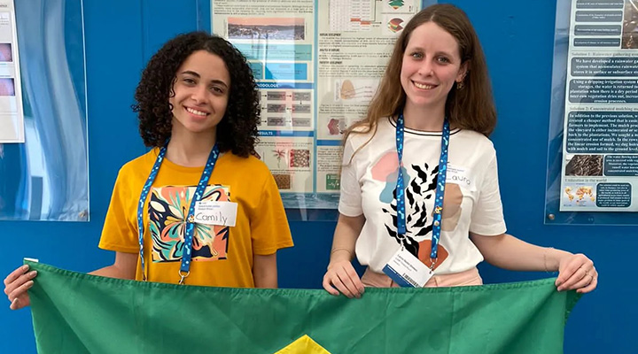 As duas garotas estão sorrindo para a foto enquanto seguram, juntas, uma bandeira do Brasil