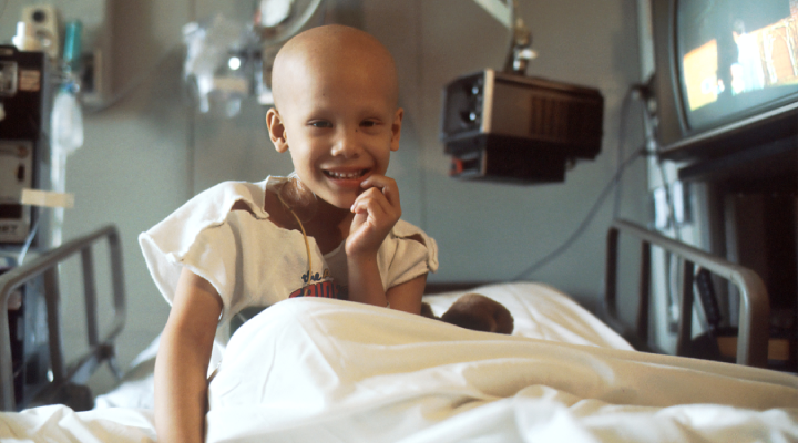 Criança com câncer sorri sentada em uma cama hospitalar