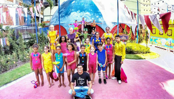 Foto de crianças e jovens vestidos com roupas coloridas. Algumas equilibram pratos em varetas, outras estão ao fundo montando uma pirâmide humana, à frente de uma lona de circo.