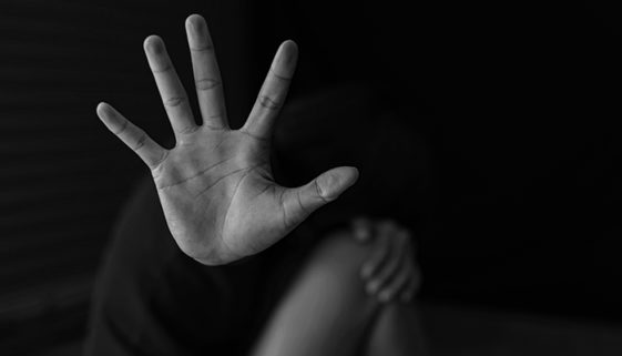 Imagem em preto e branco de mulher sentada no chão, segurando os joelhos com a mão esquerda e mostrando a palma da mão direita para a foto, no sentido de 