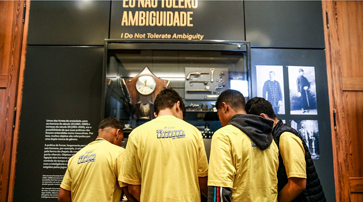 Jovens vestidos com camisetas amarelas, de costas para a foto, interagindo com expositor em museu.