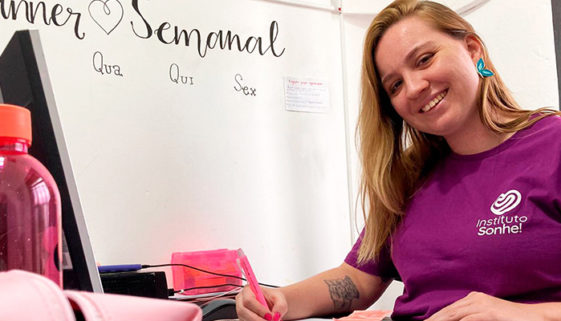 Imagem de moça sorrindo e segurando uma caneta, sentada à frente de monitor de computador. Ela veste uma camiseta de manga curta de cor rosa, com logo e nome do 