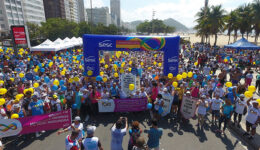 Foto panorâmica, tirada por cima, de dezenas de pessoas em volta de arco de largada da Caminhada Sesc pela Valorização da Pessoa Idosa e carregando bexigas azuis e amarelas.