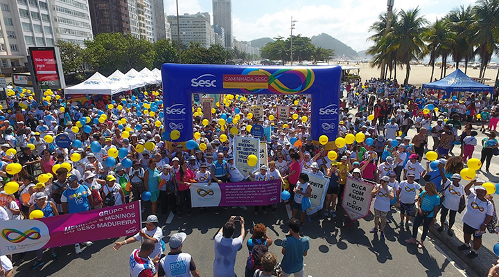 Foto panorâmica, tirada por cima, de dezenas de pessoas em volta de arco de largada da Caminhada Sesc pela Valorização da Pessoa Idosa e carregando bexigas azuis e amarelas.