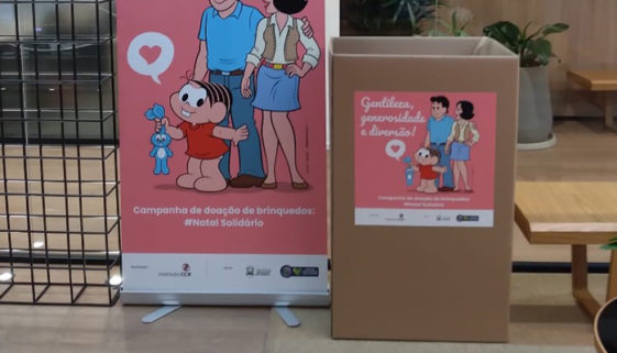 Foto de caixa de papelão e painel dispostos em via pública e estampados com desenho de Mauricio de Souza, da Mônica e seus pais, para a campanha de arrecadação de brinquedos Natal Solidário.
