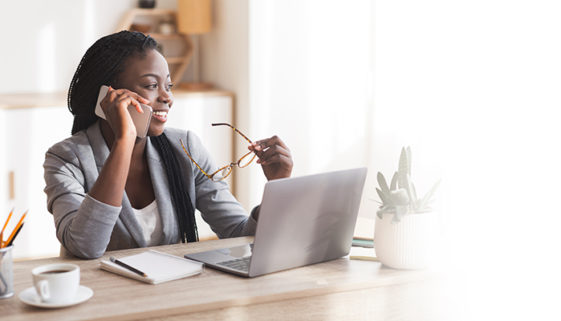 Empresária negra sorridente falando no celular no escritório moderno, sentada na mesa com óculos na mão.