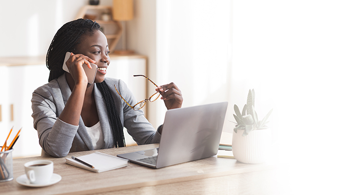 Empresária negra sorridente falando no celular no escritório moderno, sentada na mesa com óculos na mão.