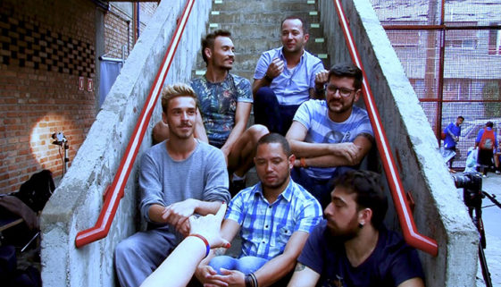 Foto de seis homens sentados nos degraus de uma escada com laterais de concreto e corrimão vermelhos.