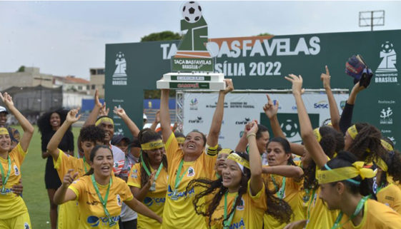 Foto de time de futebol feminino campeão. As jogadoras vestem uniforme amarelo e comemoram vitória de campeonato. Uma moça no centro de todas levanta um troféu enquanto sorri olhando pra cima.