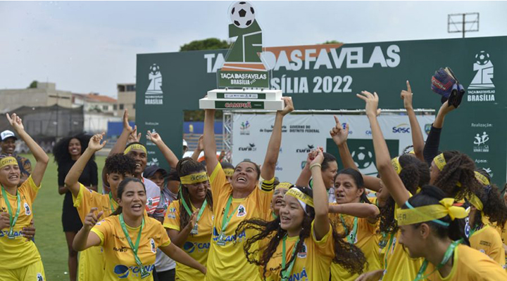 Foto de time de futebol feminino campeão. As jogadoras vestem uniforme amarelo e comemoram vitória de campeonato. Uma moça no centro de todas levanta um troféu enquanto sorri olhando pra cima.