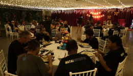 Foto de sete pessoas sentadas em cadeiras brancas, em volta de uma mesa redonda, comendo. Ao fundo, demais pessoas também estão sentadas em mesas iguais, dispostas em um salão com luzes e enfeites natalinos.